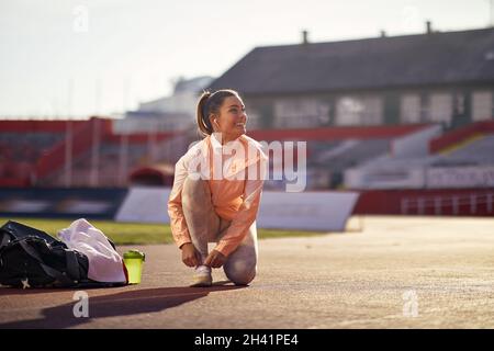 Schöne junge Erwachsene kaukasische Frau, die auf einem Knie auf der athletischen Bahn kniet, Schnürsenkel bindet, lächelt und mit ihren Kopfhörern im Ohr aufschaut Stockfoto