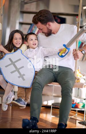 Ein junger Vater sitzt in spielerischer Atmosphäre zu Hause auf der Schaukel, während er mit den Kindern spielt. Familie, Zuhause, Freizeit Stockfoto