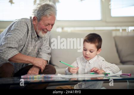 Ein kleiner Enkel liebt es zu zeichnen, während er eine gute Zeit mit seinem Opa in einer entspannten Atmosphäre zu Hause verbringt. Familie, Zuhause, Freizeit Stockfoto