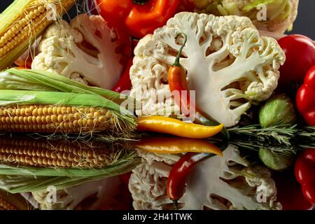 Verschiedene frische Gemüse auf einem schwarzen reflektierenden Hintergrund. Tomaten, Paprika, Blumenkohl, Chili, Gurken, Mais und Rosmarin werden präsentiert. Stockfoto