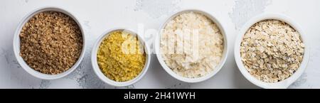 Verschiedene Getreideflocken, Reis, Hirse, Buchweizen, Haferflocken. Superfood in weißen Keramikschalen auf hellem Holzhintergrund. Draufsicht. Stockfoto