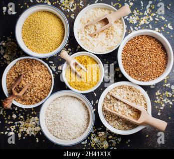 Verschiedene Getreideflocken, Reis, Hirse, Buchweizen, Haferflocken. Superfood in weißen Keramikschalen auf dunklem alten Betongrund. Draufsicht. Stockfoto