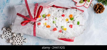 Vollkornstollen mit Rosinen und Puderzucker auf einer Leinenserviette mit Sieb, rotes Band über dem hellblau verschneiten Betonhintergrund. Tradition Stockfoto