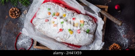 Vollkornstollen mit Rosinen und Puderzucker auf einer Leinenserviette mit Sieb, rotes Band über dem alten dunklen Betonboden. Traditionelle Germa Stockfoto