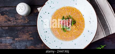 Hausgemachte rote Linsensuppe mit Speck und Rucola auf einem weißen Teller auf einem alten Holzboden. Rustikaler Stil. Draufsicht. Stockfoto