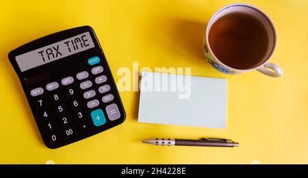 STEUERFREIER Text auf einem Rechner auf einem Desktop, Draufsicht. Eine Tasse Tee, ein Aufkleber zum Schreiben und ein Stift liegen auf einem gelben Hintergrund Stockfoto