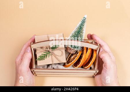 Frau Hände halten kleine Holzkiste mit Weihnachtsdekorationen - dekorative Weihnachtsbaum, trockene Orangenscheiben, DIY verpackt Geschenkbox auf beigem Hintergrund. H Stockfoto