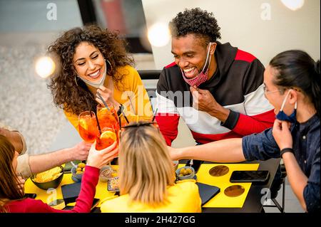 Glückliche multiethnische Freunde, die ihre Masken absenken, lächeln und stoßen im Cocktailbargarten mit Spritz an - Freundschaftskonzept mit jungen Leuten, die Fu haben Stockfoto