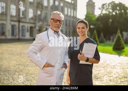 Professioneller leitender männlicher Arzt in Laborkittel und -Brille, der die Kamera anschaut und zusammen mit einer lächelnden jungen Krankenschwester im Freien in der Nähe der Klinik posiert Stockfoto