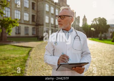 Seriöser leitender Arzt im Laborkittel und in einer Brille, die die Zwischenablage hält, zur Seite schaut und in der Nähe der Klinik im Freien steht Stockfoto