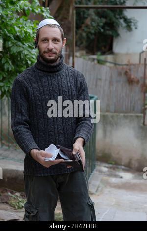 Mann zeigt leere Brieftasche. Jüdischer Mann mit Kippah auf Hebräisch oder Yarmulke mit grünen Bäumen im Hintergrund, Bart mit schwarzem Sweatshirt. Kein Geld Stockfoto