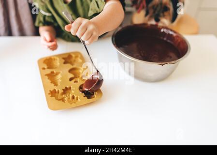Kinderhände gießen Schokoladenteig in eine Silikonform auf einem weißen Tisch. Weihnachts-Lebkuchen. Stockfoto