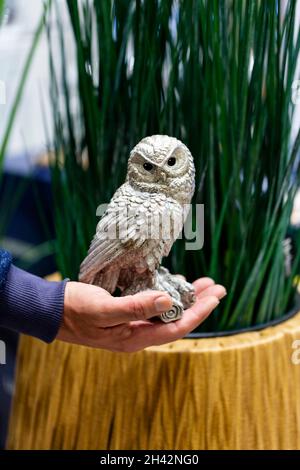 Eule Spielzeug Figur mit der Hand halten, Nahaufnahme. Eine Hand, die eine Eule auf unscharfem grünen Hintergrund hält, selektiver Fokus. Gartenskulptur, Keramik, kinderl Stockfoto
