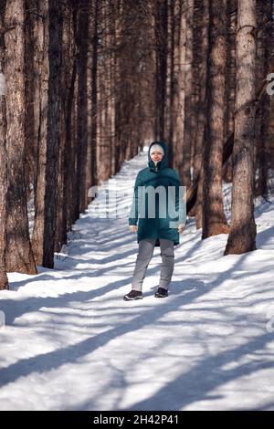 Kaukasische Frau im verschneiten Wald. Junge attraktive Frau in einer warmen Daunenjacke spazieren auf einer mit Schnee bedeckten Landstraße nach dem Schneesturm Stockfoto