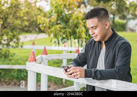 Unternehmungslustiger junger Mann neben dem weißen Holzzaun mit seinem Handy in den Händen, der seine sozialen Netzwerke anschaut, glücklich über seine Leistungen, Witz Stockfoto