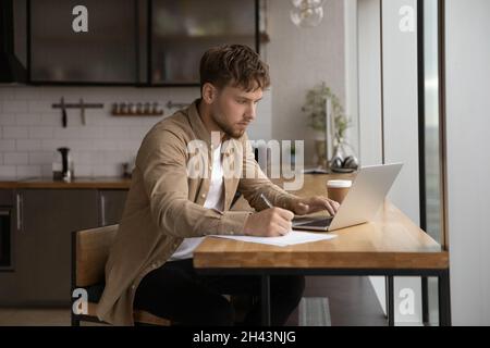 Konzentrierter junger Mann, der am Computer arbeitet und Notizen schreibt. Stockfoto