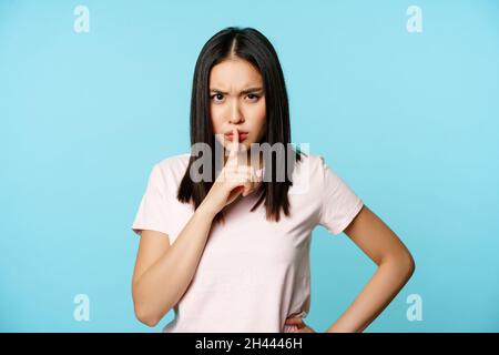 Wütende asiatische Frau huschend, vor Missbilligung runzelte Stirn und huschte, Tabuzeichen, Stille-Geste, auf blauem Hintergrund stehend. Stockfoto