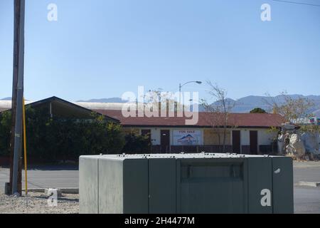 Kleiner Versorgungsschuppen oder Servicehaus mit abgeschrägtem Dach und Elektrokasten, Versorgungsmast in der Nähe in offener, trockener Wüstenlandschaft unter klarem blauem Himmel, ru Stockfoto