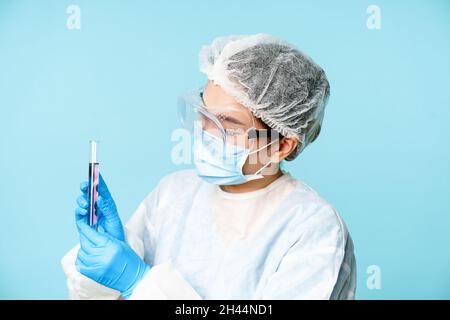 Labormitarbeiter, der Chemikalien im Probenröhrchen mischt, persönliche Schutzausrüstung und Schutzbrille trägt, blauer Hintergrund Stockfoto