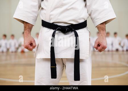 Karate-Meister in einem weißen Kimono und mit einem schwarzen Gürtel, steht vor der Formation seiner Schüler. Kampfkunstschule in der Turnhalle trainieren Stockfoto