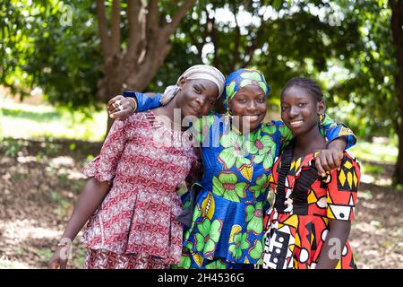 Drei strahlende junge afrikanische Frauen in schönen traditionellen Kleidern, die die Kamera betrachten; Konzept der menschlichen Beziehung Stockfoto