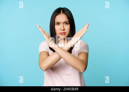 Stopp. Besorgte asiatische Frau, die ein Kreuzzeichen zeigt, nein sagt, Bewusstsein wecken, auf blauem Hintergrund stehend Stockfoto