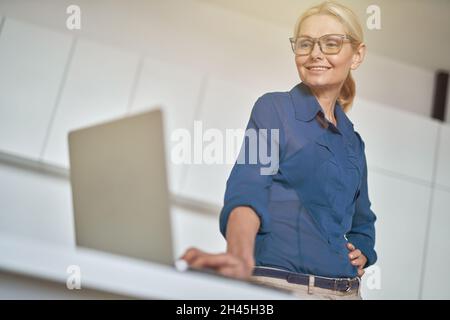 Porträt einer erfolgreichen reifen Geschäftsfrau in einer lächelnden Brille, die am Laptop arbeitet, in der modernen Küche zu Hause steht Stockfoto