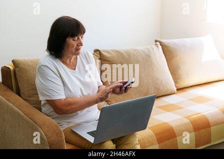 Ältere Frau mit Laptop-Computer sitzt auf der Couch und spricht mit jemandem am Telefon. Neue Technologien sind einfacher und schneller zu verwenden. Aufnahme einer schönen älteren Frau, die am Telefon anruft und einen Laptop benutzt, Stockfoto