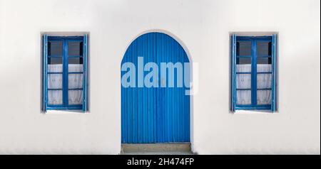 Blaue Vintage-Tür mit Bogenfenstern mit geöffneten Fensterläden und Vorhängen auf einem leeren weiß getünchten Wandhintergrund. Traditionelle griechische Insel Stockfoto