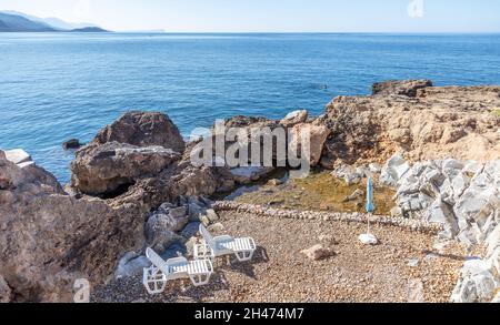 Sommerurlaub in Griechenland neben Ägäis blau ruhigen Meer Hintergrund. Menschenleerer, steiniger Strand mit zwei Liegen und einem Sonnenschirm. Weißer Plastik Strand ch Stockfoto