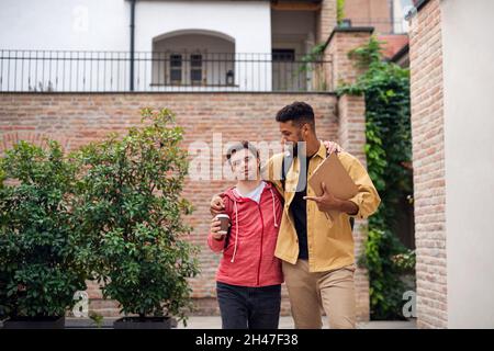 Junger Mann mit Down-Syndrom und sein Mentoring-Freund gehen und umarmen im Freien Stockfoto