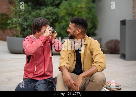 Glücklicher junger Mann mit Down-Syndrom sitzt und fotografiert mit der Kamera eines Freundes im Freien Stockfoto