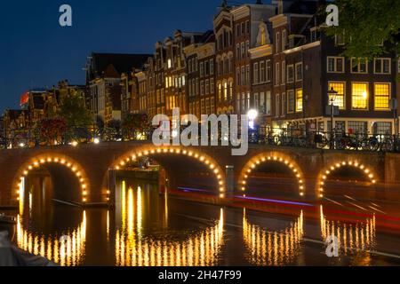 Niederlande. Nacht auf dem Amsterdamer Kanal. Beleuchtete Brücke und Bootspfad. Traditionelle holländische Häuser am Wasser Stockfoto