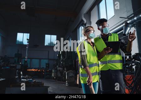 Industrieinspektoren mit Gesichtsmasken, die in Innenräumen in der Metallwerkstatt eine allgemeine Überprüfung durchführen, Coronavirus-Konzept. Stockfoto