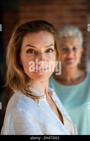 Junge Frau, die die Kamera anschaut, ihre Mutter im Hintergrund, die sie anschaut. Stockfoto