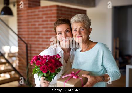 Glückliche ältere Mutter bekommt Geschenk und Blumenstrauß von erwachsenen Tochter drinnen zu Hause, Blick auf die Kamera. Stockfoto