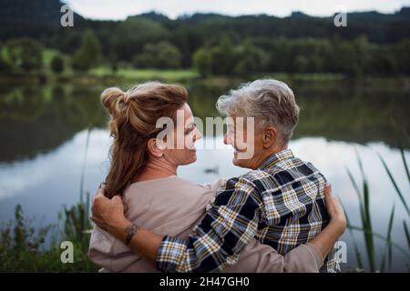 Rückansicht einer glücklichen älteren Mutter, die sich mit einer erwachsenen Tochter umarmt, wenn sie am See in der Natur sitzt Stockfoto