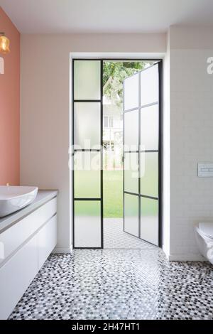 Badezimmer mit belgischer Tür zum Garten. Ein länglicher Schrank mit Waschbecken, pfirsichfarbener Wand und einer an der Wand hängenden Leuchte. Stockfoto