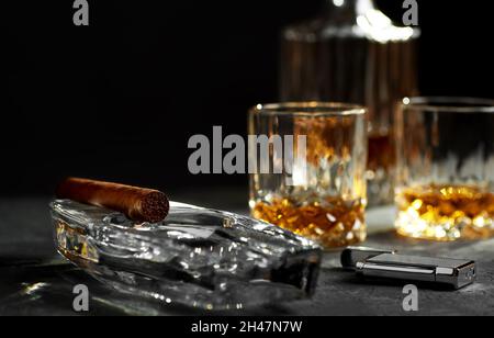 Stillleben mit einer kubanischen Zigarre in einem Aschenbecher, einem Glas und einer Flasche Whiskey und einem Feuerzeug auf einer steinernen Tischplatte. Schwarzer Hintergrund mit Platz für Text. Stockfoto