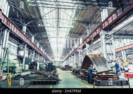 Innenraum der metallurgischen Anlage. Große Werkstatt mit Metallbearbeitungsmaschinen. Arbeiter in Masken entladen Metallbleche auf einem Kran. Stockfoto