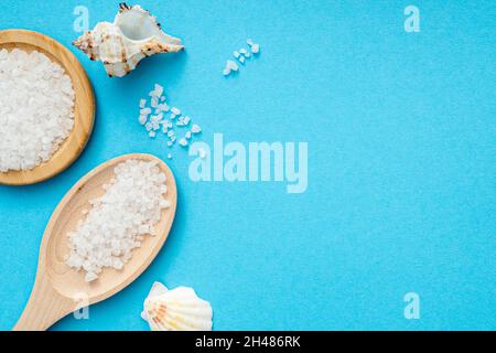 Meersalz, Holzlöffel, Muscheln auf blauer Oberfläche. Flach mit Salzkristallen, Draufsicht, Platz für Text Stockfoto