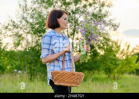 Lächelnde Frau mittleren Alters mit einem Korb und einem Blumenstrauß in der Natur Stockfoto