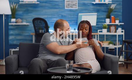 Interracial paar mit Schwangerschaft Film im Fernsehen beim Essen Popcorn. Multiethnische Eltern erwarten ein Baby und sitzen auf der Wohnzimmercouch. Gemischtes Volk, das sich mit dem Fernsehen entspannt Stockfoto