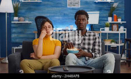 POV von interracial paar Film im Fernsehen mit Snack zu Hause. Verheiratete multiethnische Liebhaber, die beim Essen von Popcorn im Wohnzimmer auf die Kamera schauten. Gemischte Rassen Menschen kleben auf dem Sofa Stockfoto