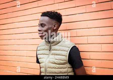 Positiver junger afroamerikanischer Tausendjähriger Mann mit trendigem Haarschnitt im T-Shirt und stilvoller Kugelweste, die neben einer Ziegelwand steht und lächelt Stockfoto