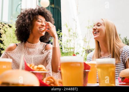 Zwei attraktive, lebhafte junge Frauen genießen Snacks und ein Bier zusammen in einer Bar, die sich mit Pommes frites füttern und sich auf eine schöne schwarze Frau konzentrieren, die lacht und Stockfoto