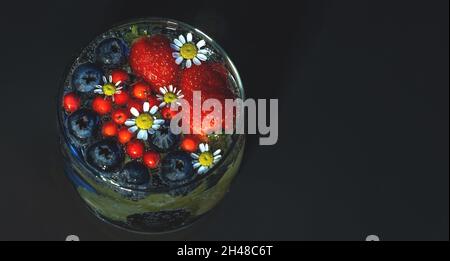 Ein Glas mit einem duftenden Getränk aus frischen Beeren und Kamille, Draufsicht. Layout mit Platz für Text, auf dunklem Hintergrund. Flach liegend, Kopierplatz. Stockfoto