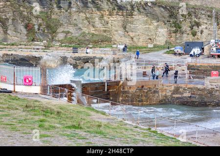 Die Menschen stehen und beobachten die Wellen, die gegen die Klippen krachen, bevor sie bei Flut in Portreath, Cornwall, den Hafen betreten Stockfoto