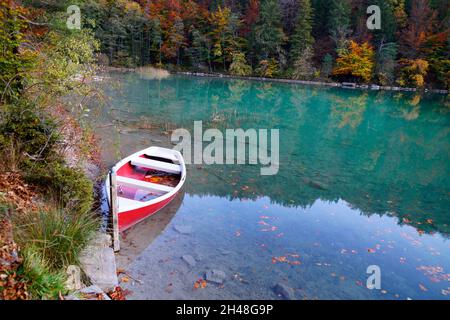 Eine malerische Aussicht auf den Alatsee und ein rotes Boot, das an einem schönen Herbsttag auf dem smaragdgrünen Wasser ruht (Bayern in Deutschland) Stockfoto