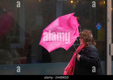 Starker Wind und Frau, die einen umgedrehten rosa Regenschirm in Preston, Lancashire, Großbritannien, trägt. 1st. November 2021. UK Wetter, kaputter Regenschirm, invertiert, innen heraus, verbogener gebrochener Wind und Regen. Geschäfte, Einkäufer, die an einem nassen und windigen Tag im Stadtzentrum von Preston einkaufen. Quelle: MediaWorlImages/AlamyLiveNews Stockfoto